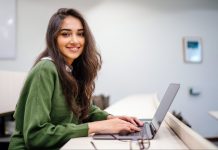 Junge Frau an einem Schreibtisch blickt seitlich vom Laptop auf.