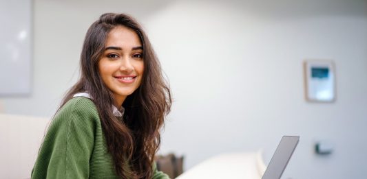 Junge Frau an einem Schreibtisch blickt seitlich vom Laptop auf.
