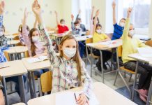 Kinder mit Masken in einem Klassenraum melden sich.