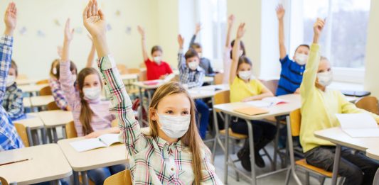 Kinder mit Masken in einem Klassenraum melden sich.