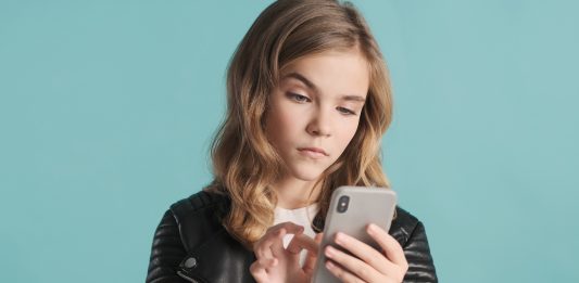 langhaariges Mädchen vor blauem Hintergrund tippt mit skeptischem Blick auf ihr Smartphone.