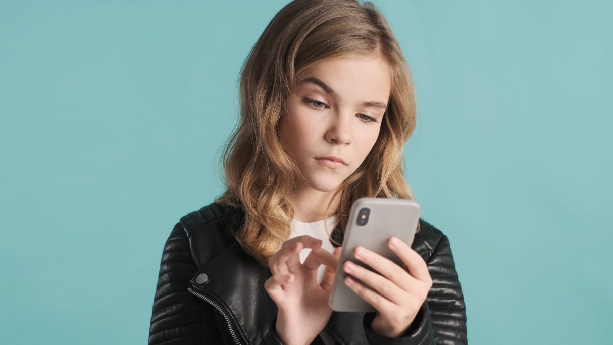 langhaariges Mädchen vor blauem Hintergrund tippt mit skeptischem Blick auf ihr Smartphone.