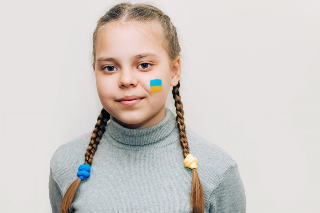 bezopftes Mädchen in grauem Pullover, mit ukrainischer Flagge auf der Wange