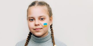 bezopftes Mädchen in grauem Pullover, mit ukrainischer Flagge auf der Wange