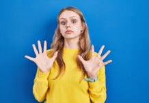 junge blonde Frau in gelbem Pullover mit ablehnender Geste vor blauem Hintergrund