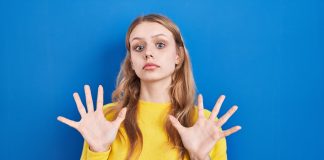 junge blonde Frau in gelbem Pullover mit ablehnender Geste vor blauem Hintergrund