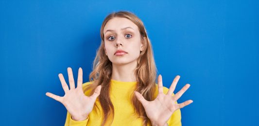 junge blonde Frau in gelbem Pullover mit ablehnender Geste vor blauem Hintergrund
