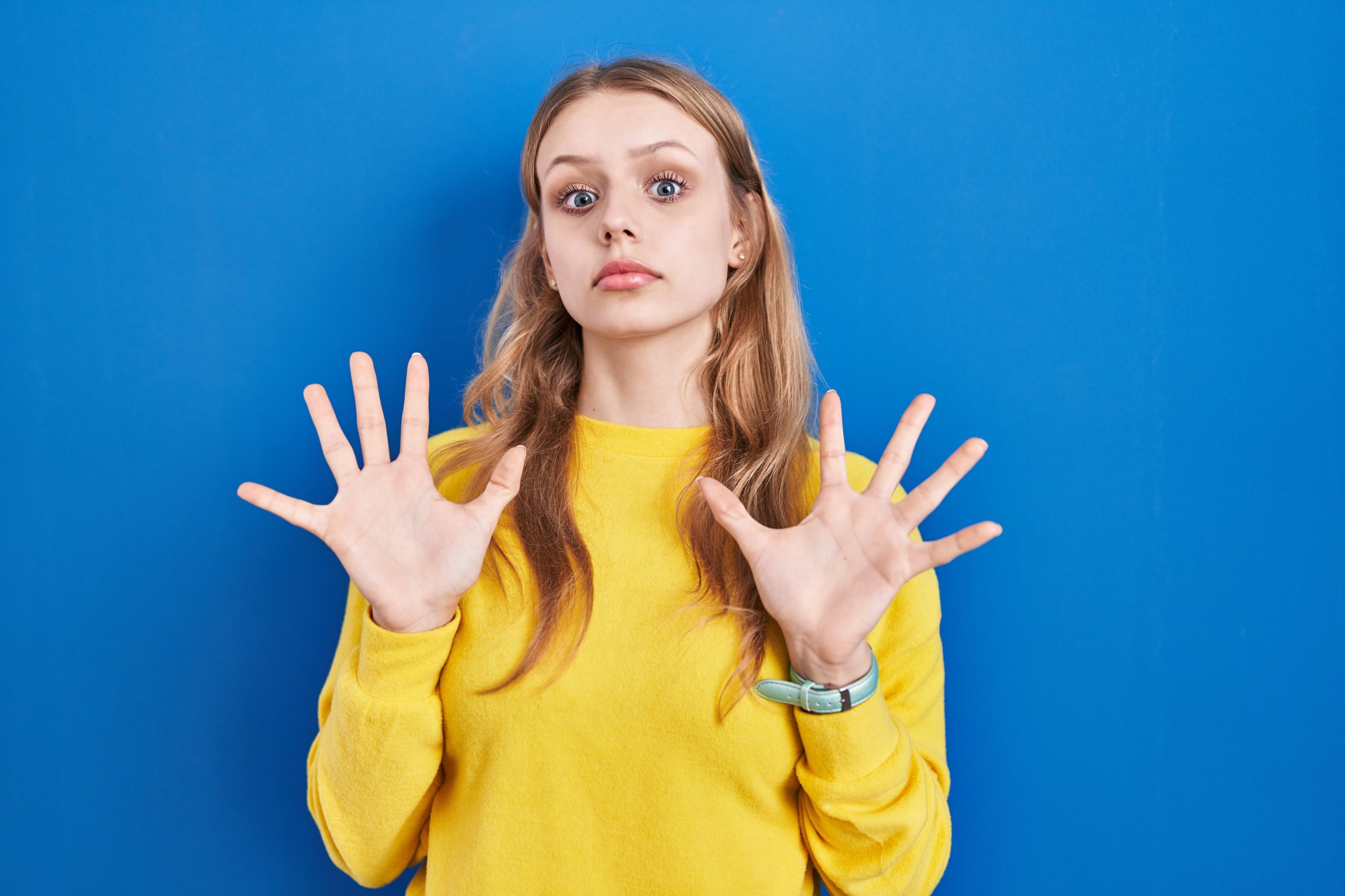 junge blonde Frau in gelbem Pullover mit ablehnender Geste vor blauem Hintergrund