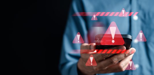 Warnzeicheneinblendungen um ein Handy, das jemand mit einem blauen Hemd vor dunklem Hintergrund in die Kamera hält.