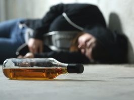 Im Vordergrund eine leere Flasche, im Hintergrund ein offenbar berauschter liegender Jugendlicher in einen Kapuzenpullover.