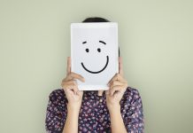 Jemand in kleingemustertem Kleid hält sich eine lächelnde Punkt-Punkt-Komma-Strich-Zeichnung vor das Gesicht.