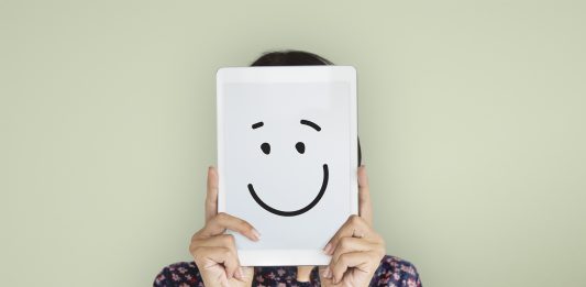 Jemand in kleingemustertem Kleid hält sich eine lächelnde Punkt-Punkt-Komma-Strich-Zeichnung vor das Gesicht.