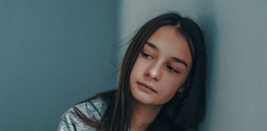 Ein an die Wand gelehntes Mädchen schaut misstrauisch zweifelnd in den Raum.